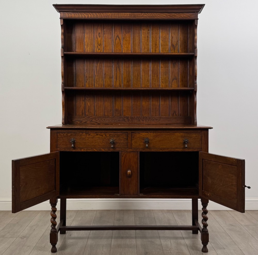 An early 20th-century dark oak Welsh dresser featuring a three-tier open plate rack mounted on a base cabinet with two drawers, geometric panel doors, and barley twist legs. View II