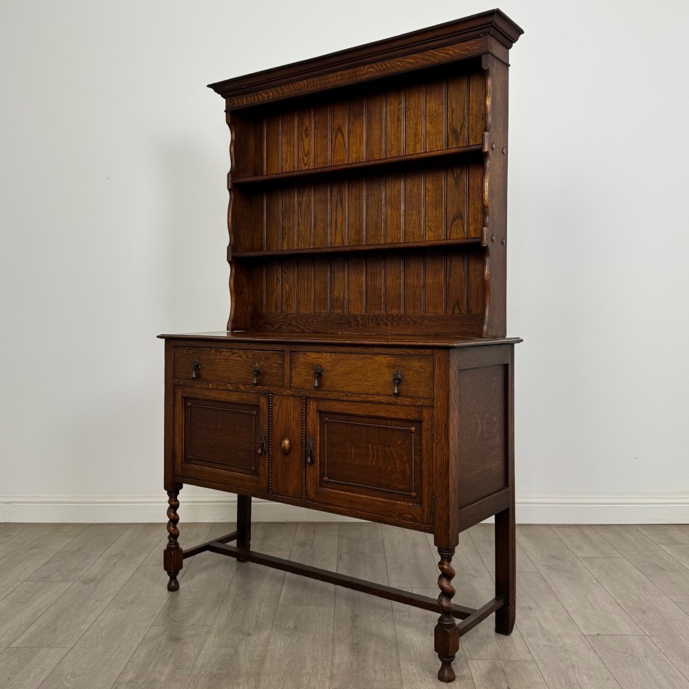 An early 20th-century dark oak Welsh dresser featuring a three-tier open plate rack mounted on a base cabinet with two drawers, geometric panel doors, and barley twist legs. View III
