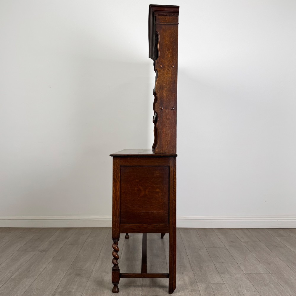 An early 20th-century dark oak Welsh dresser featuring a three-tier open plate rack mounted on a base cabinet with two drawers, geometric panel doors, and barley twist legs. View IV
