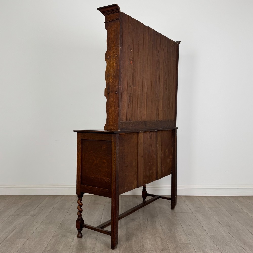 An early 20th-century dark oak Welsh dresser featuring a three-tier open plate rack mounted on a base cabinet with two drawers, geometric panel doors, and barley twist legs. View V