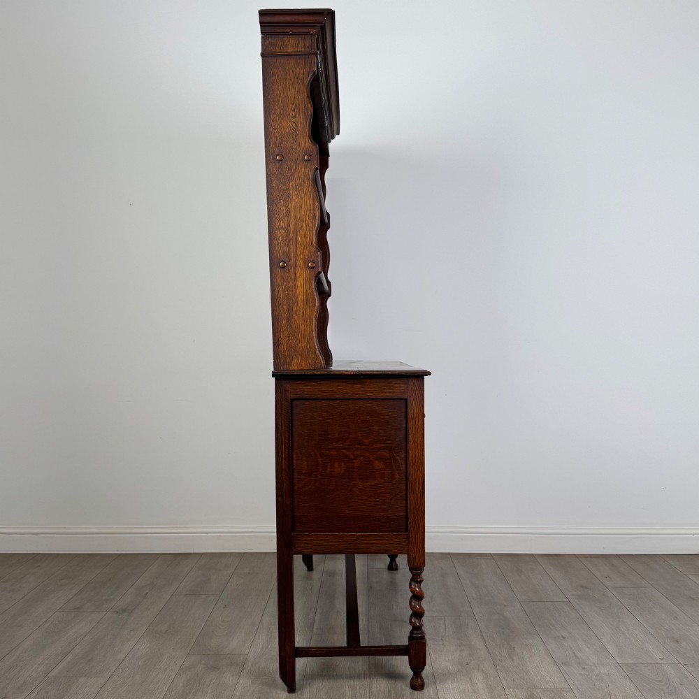 An early 20th-century dark oak Welsh dresser featuring a three-tier open plate rack mounted on a base cabinet with two drawers, geometric panel doors, and barley twist legs. View VIII
