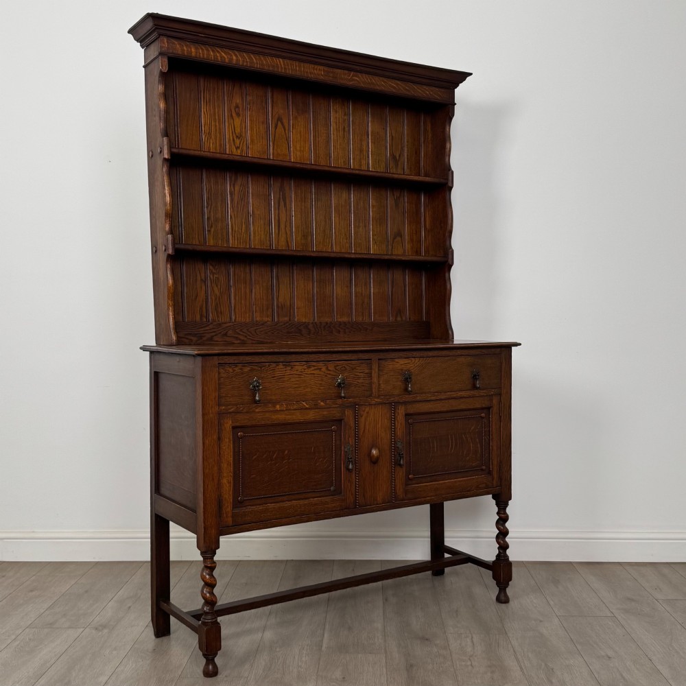 An early 20th-century dark oak Welsh dresser featuring a three-tier open plate rack mounted on a base cabinet with two drawers, geometric panel doors, and barley twist legs. View IX