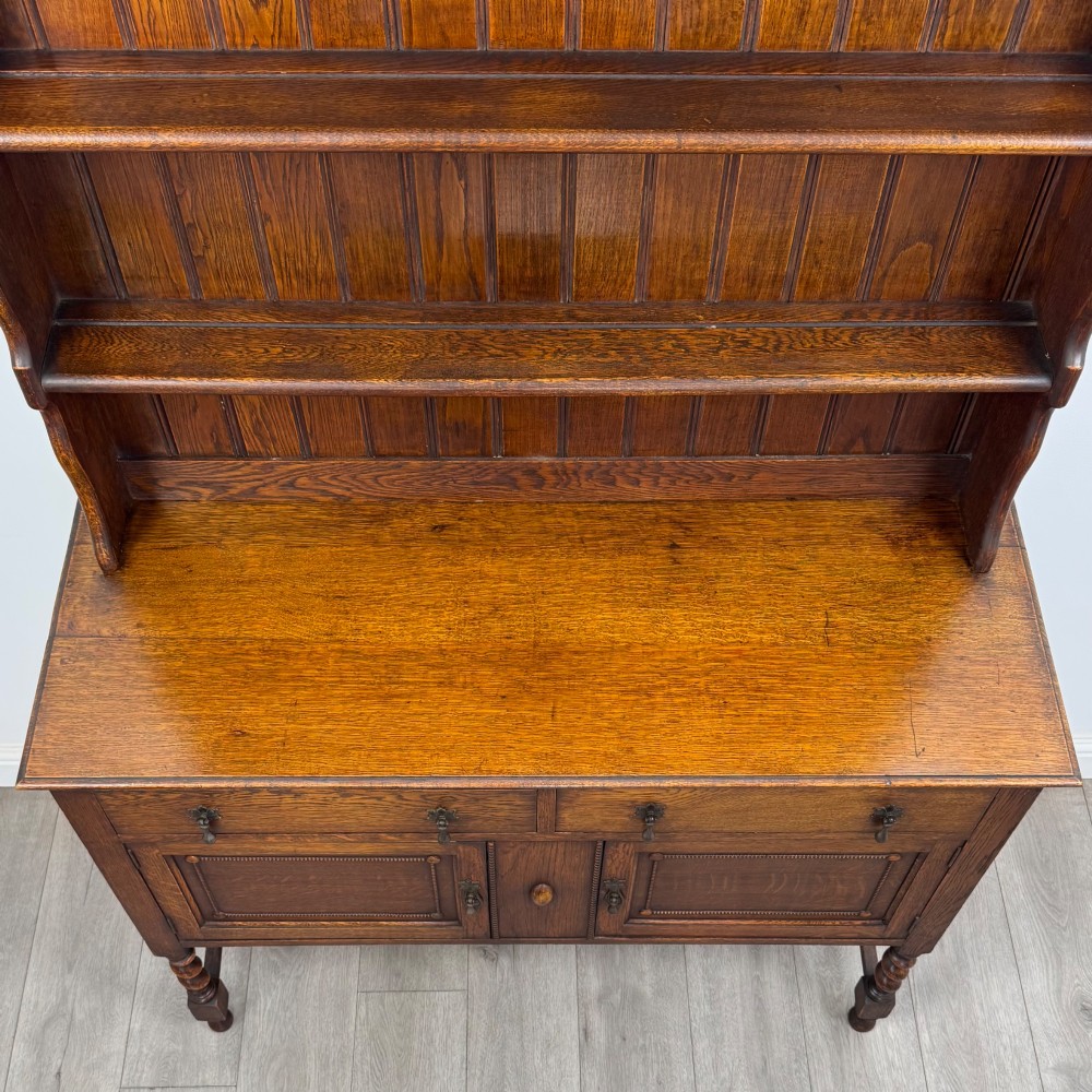 An early 20th-century dark oak Welsh dresser featuring a three-tier open plate rack mounted on a base cabinet with two drawers, geometric panel doors, and barley twist legs. View X