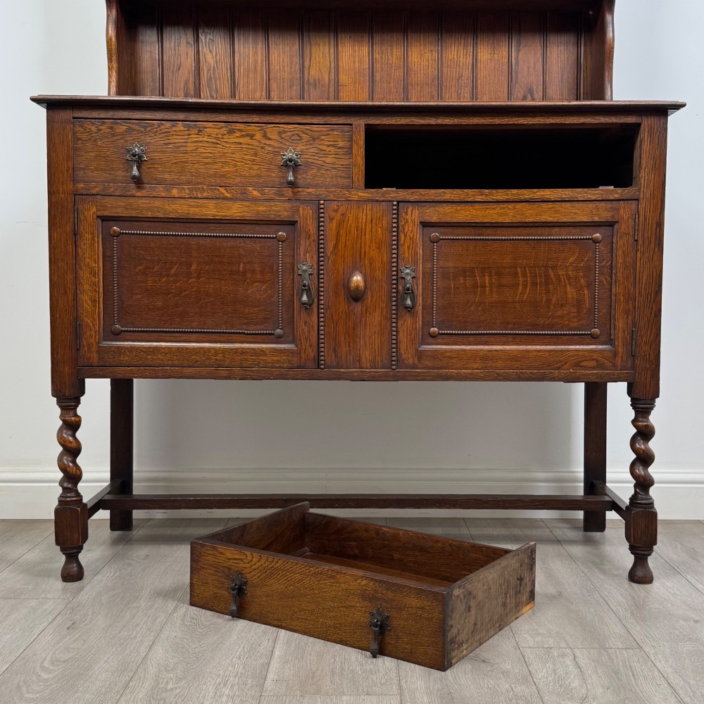 An early 20th-century dark oak Welsh dresser featuring a three-tier open plate rack mounted on a base cabinet with two drawers, geometric panel doors, and barley twist legs. View XI