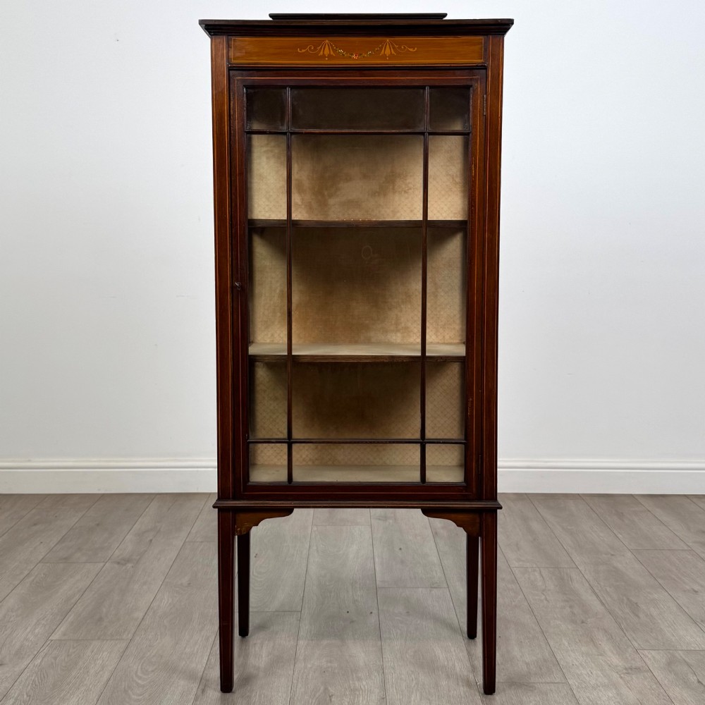 A slender Edwardian mahogany display cabinet featuring a single glazed door with geometric glazing bars, floral inlay on the top frieze, and long tapered legs.