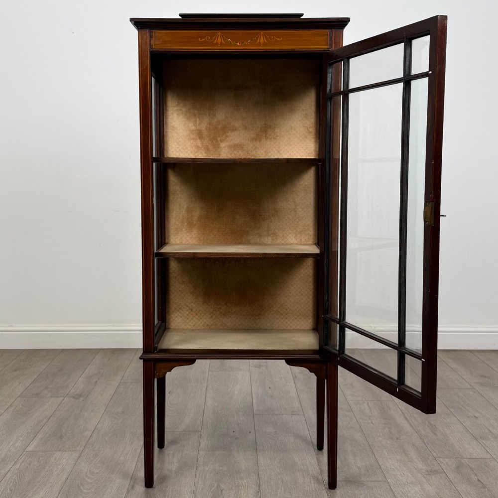 A slender Edwardian mahogany display cabinet featuring a single glazed door with geometric glazing bars, floral inlay on the top frieze, and long tapered legs. View II