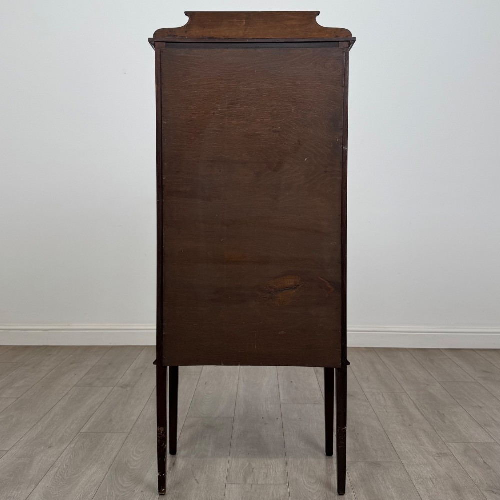 A slender Edwardian mahogany display cabinet featuring a single glazed door with geometric glazing bars, floral inlay on the top frieze, and long tapered legs. View VI