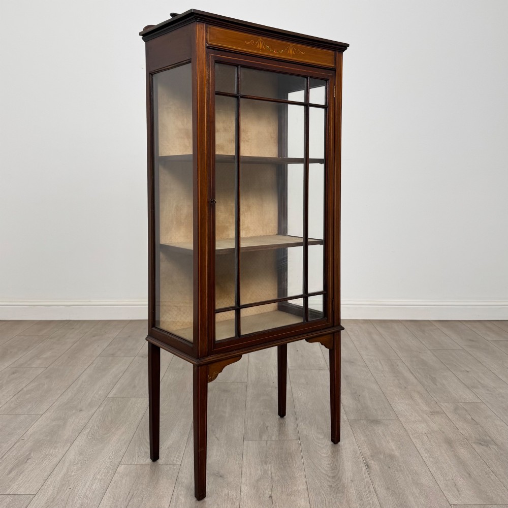 A slender Edwardian mahogany display cabinet featuring a single glazed door with geometric glazing bars, floral inlay on the top frieze, and long tapered legs. View IX
