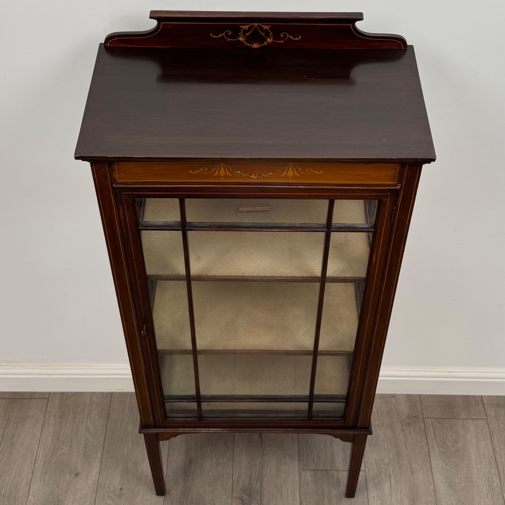 A slender Edwardian mahogany display cabinet featuring a single glazed door with geometric glazing bars, floral inlay on the top frieze, and long tapered legs. View X
