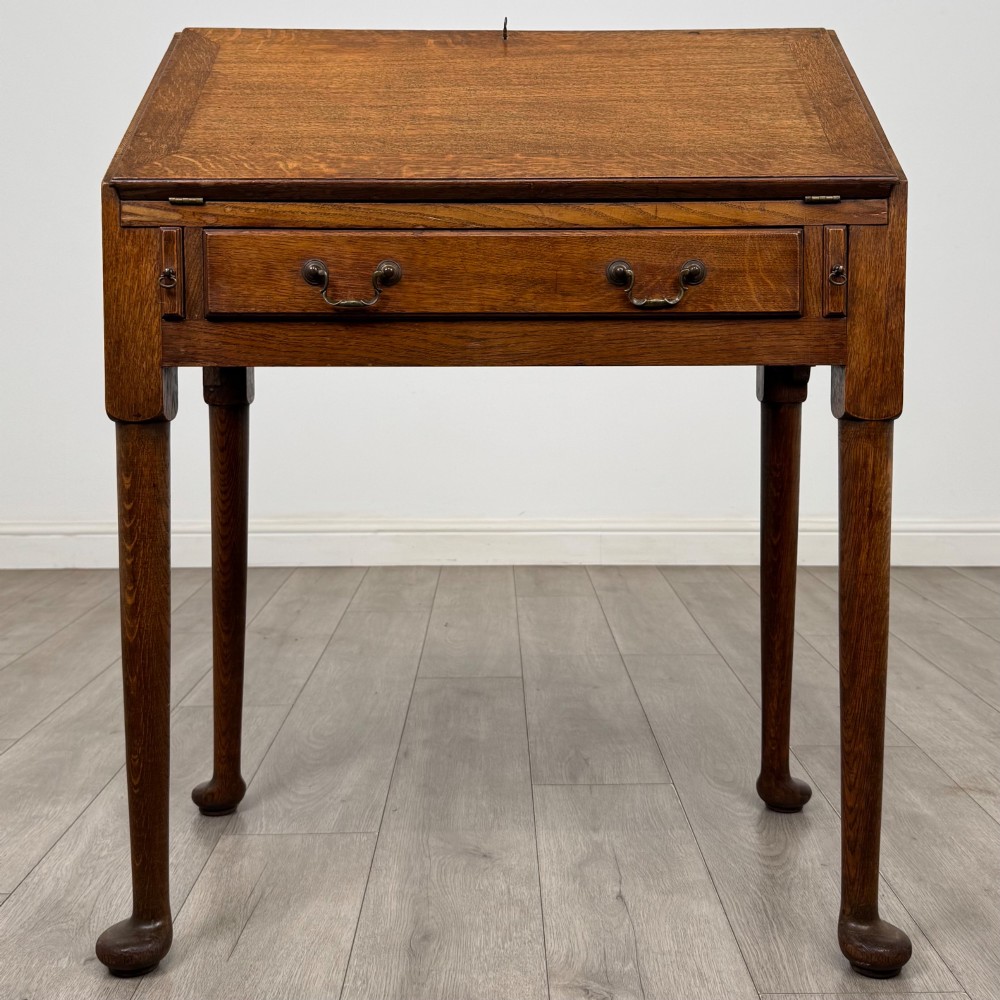 A golden oak slant-front writing bureau standing on four slender legs with pad feet, featuring a single lower drawer with drop handles and a hinged writing slope.