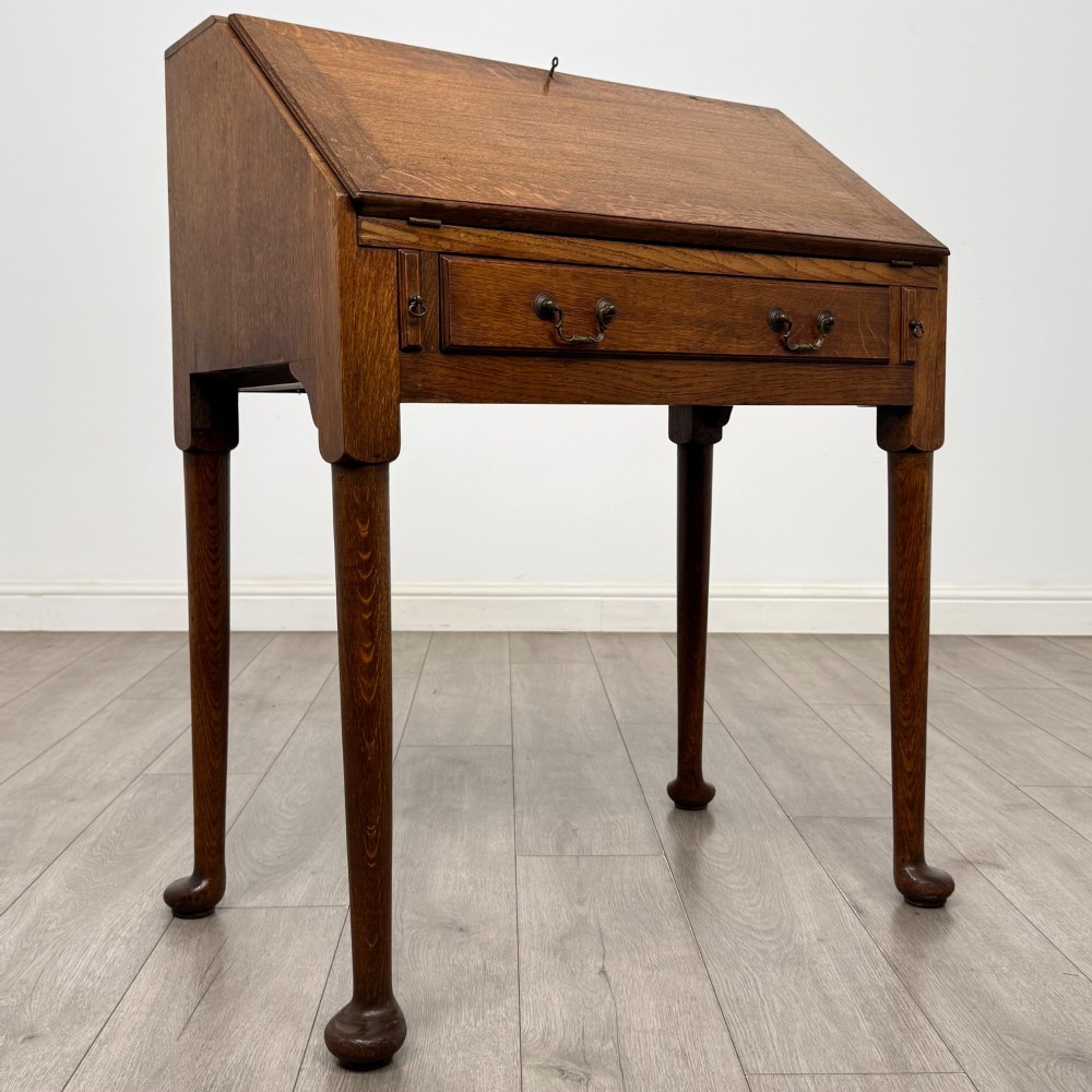 A golden oak slant-front writing bureau standing on four slender legs with pad feet, featuring a single lower drawer with drop handles and a hinged writing slope. View IX