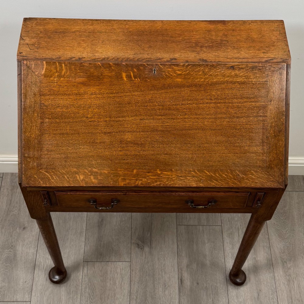 A golden oak slant-front writing bureau standing on four slender legs with pad feet, featuring a single lower drawer with drop handles and a hinged writing slope. View X