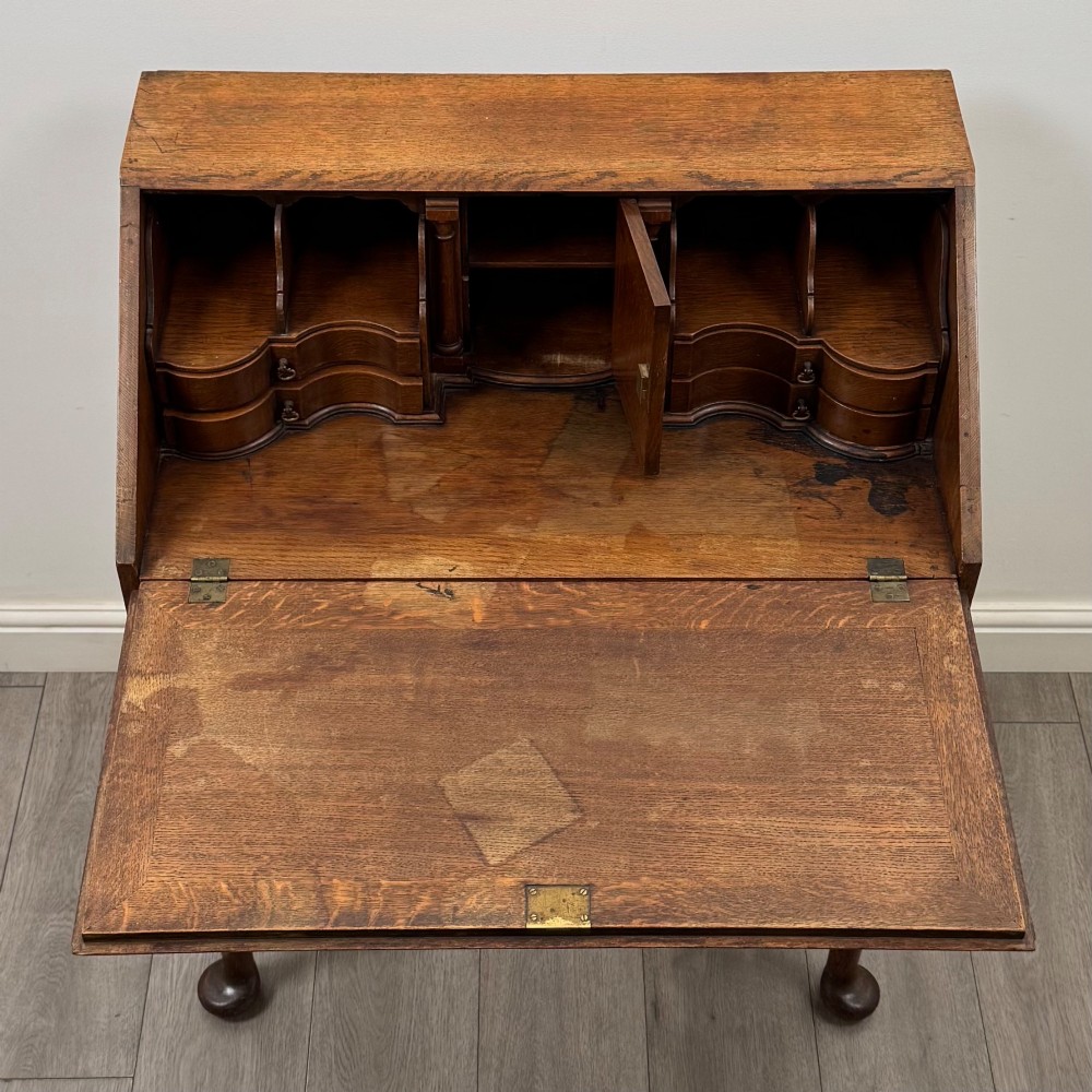 A golden oak slant-front writing bureau standing on four slender legs with pad feet, featuring a single lower drawer with drop handles and a hinged writing slope. View XI