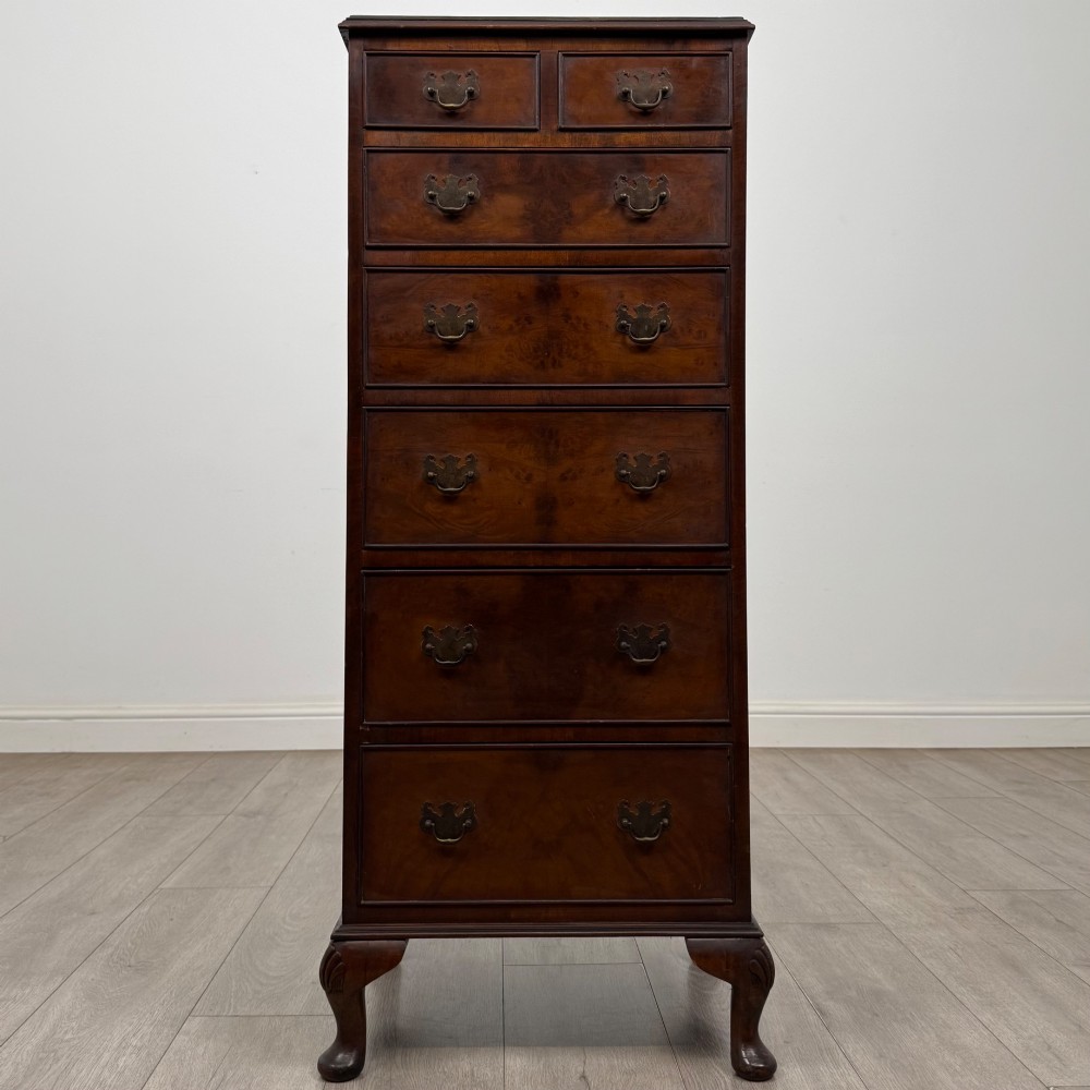A tall, narrow dark brown burr walnut chest of drawers featuring brass handles and carved cabriole legs.