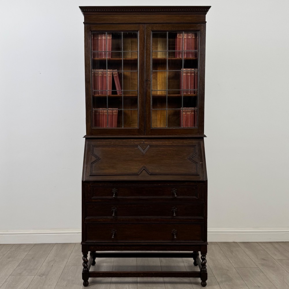 A tall 20th-century Jacobean Revival dark oak bureau bookcase with a leaded glass cabinet above a slant-front writing desk and three lower drawers.