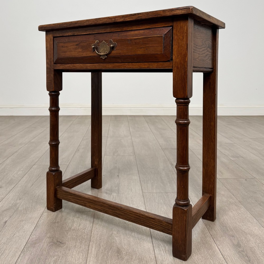 A small dark oak side table featuring a single drawer with a brass handle, turned front legs, and lower stretchers. View II