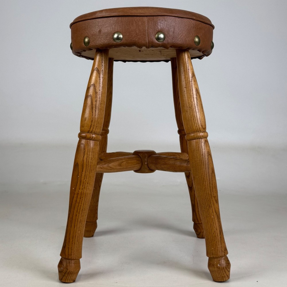A circular oak stool featuring a tan brown leather padded seat secured with brass studs and supported by four turned legs with stretchers. View II