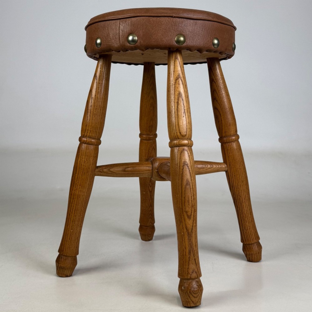 A circular oak stool featuring a tan brown leather padded seat secured with brass studs and supported by four turned legs with stretchers. View III