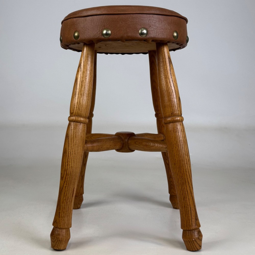 A circular oak stool featuring a tan brown leather padded seat secured with brass studs and supported by four turned legs with stretchers. View IV