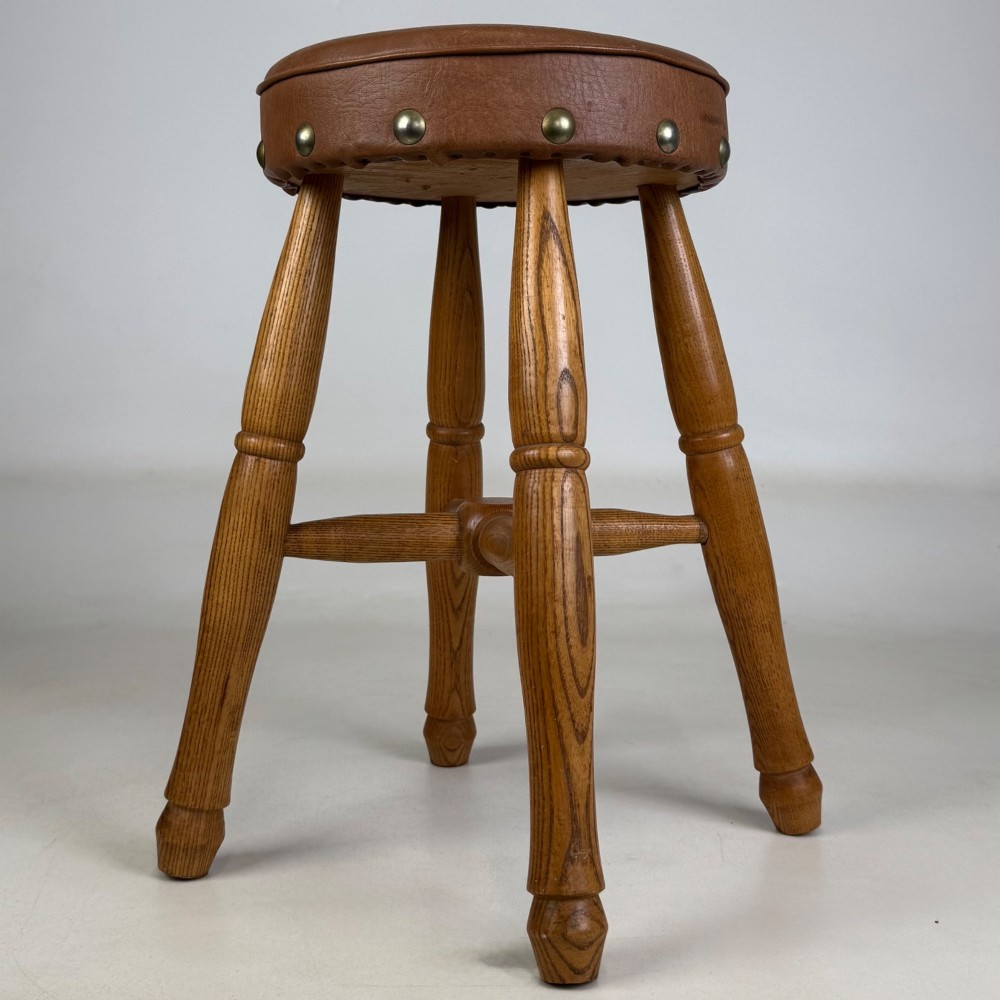 A circular oak stool featuring a tan brown leather padded seat secured with brass studs and supported by four turned legs with stretchers. View V