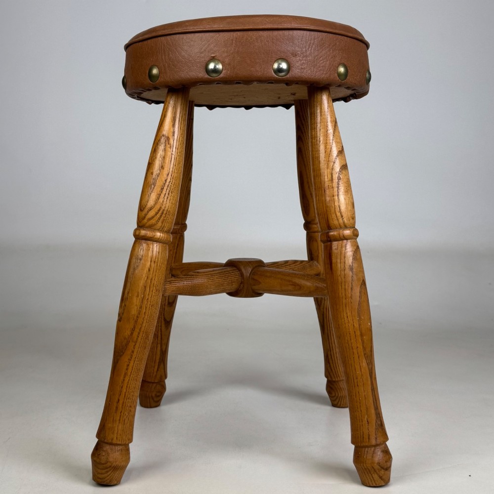 A circular oak stool featuring a tan brown leather padded seat secured with brass studs and supported by four turned legs with stretchers. View VI