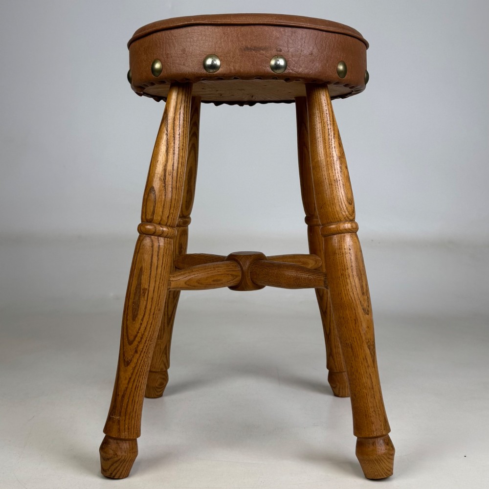 A circular oak stool featuring a tan brown leather padded seat secured with brass studs and supported by four turned legs with stretchers. View VII