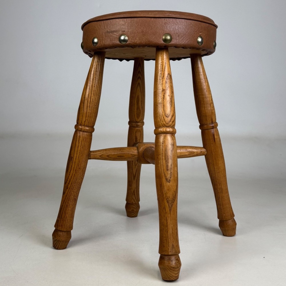 A circular oak stool featuring a tan brown leather padded seat secured with brass studs and supported by four turned legs with stretchers. View VIII