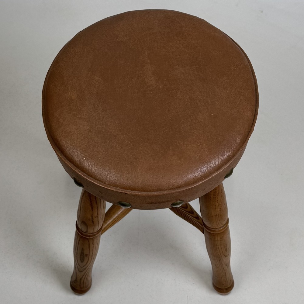 A circular oak stool featuring a tan brown leather padded seat secured with brass studs and supported by four turned legs with stretchers. View IX
