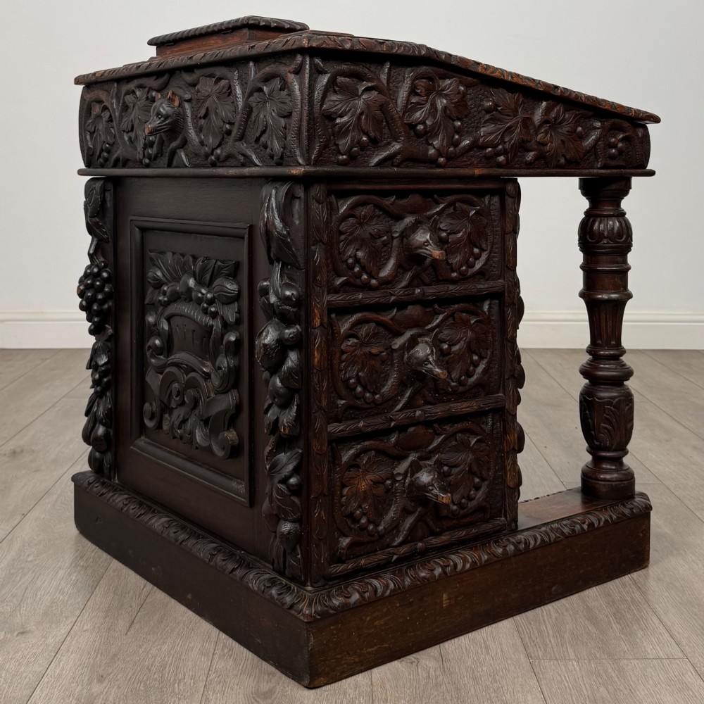 A 19th-century Victorian carved oak davenport desk featuring heavy foliate carvings, turned columns, and a green leather insert. View VI