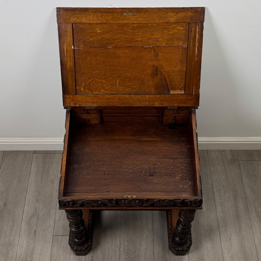 A 19th-century Victorian carved oak davenport desk featuring heavy foliate carvings, turned columns, and a green leather insert. View X