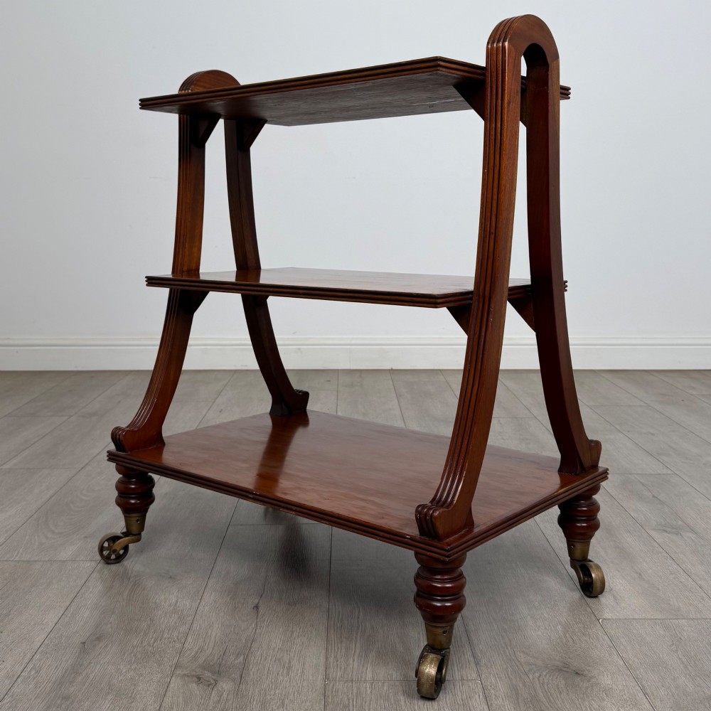 A three-tiered mahogany trolley featuring arched reeded side supports and turned feet resting on brass castors.