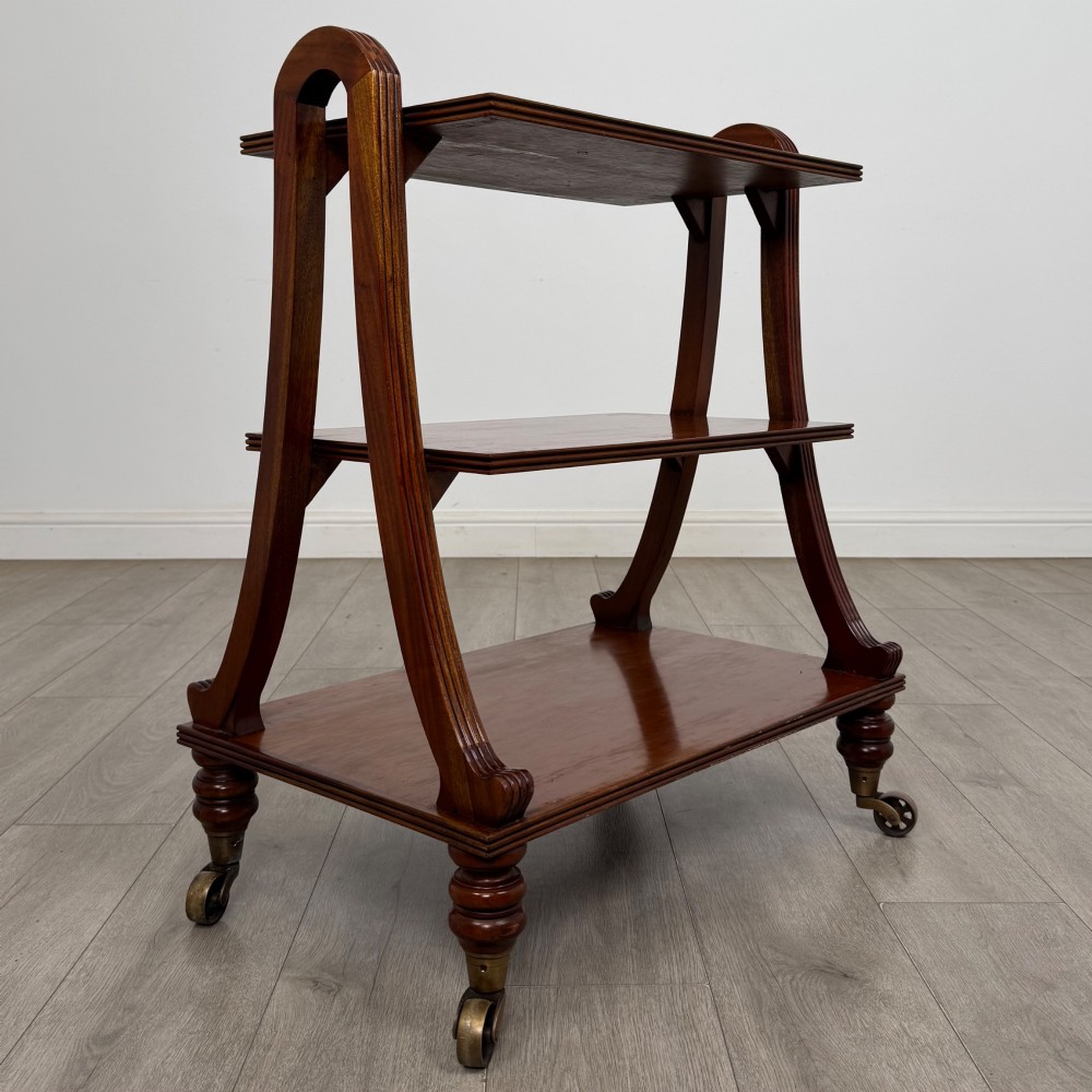 A three-tiered mahogany trolley featuring arched reeded side supports and turned feet resting on brass castors. View III
