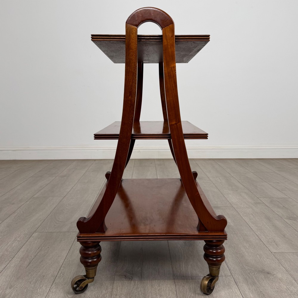 A three-tiered mahogany trolley featuring arched reeded side supports and turned feet resting on brass castors. View VI