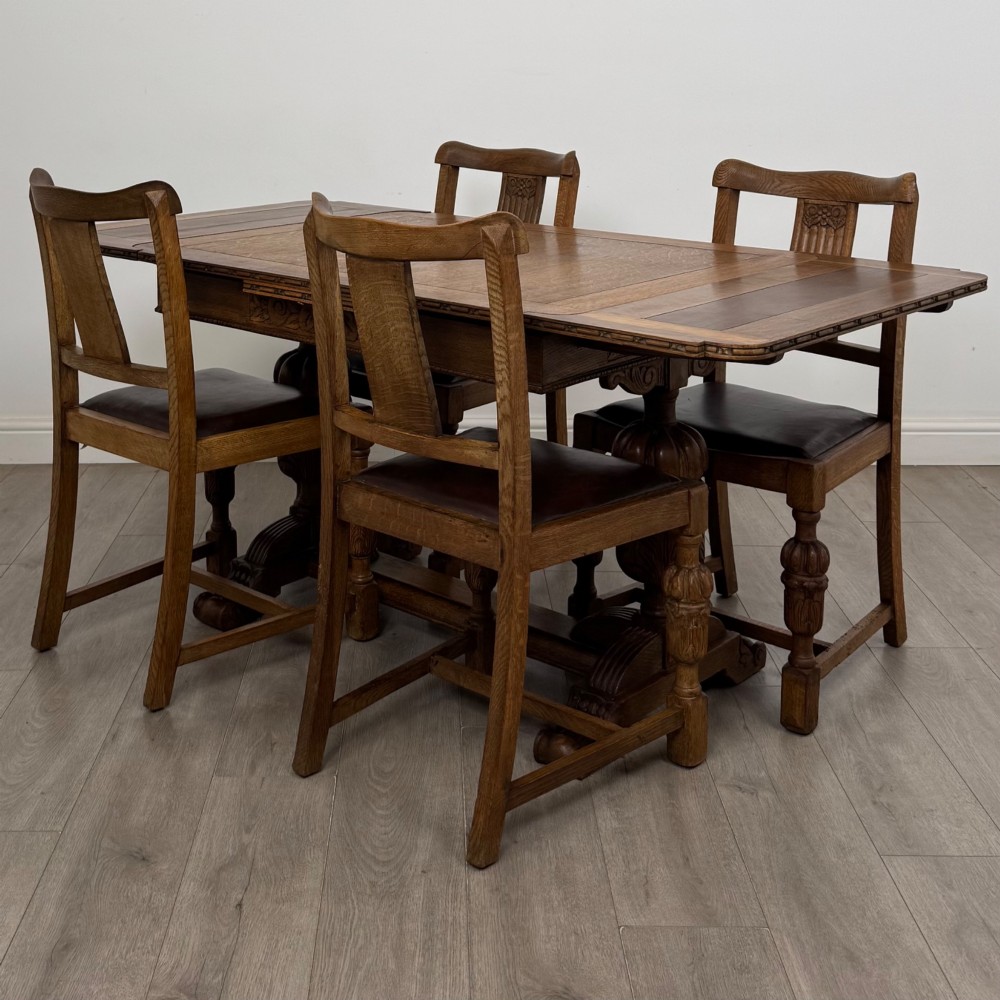 An early twentieth-century oak dining table featuring carved frieze detailing and heavy turned legs, shown alongside four matching oak dining chairs with dark leather seats.