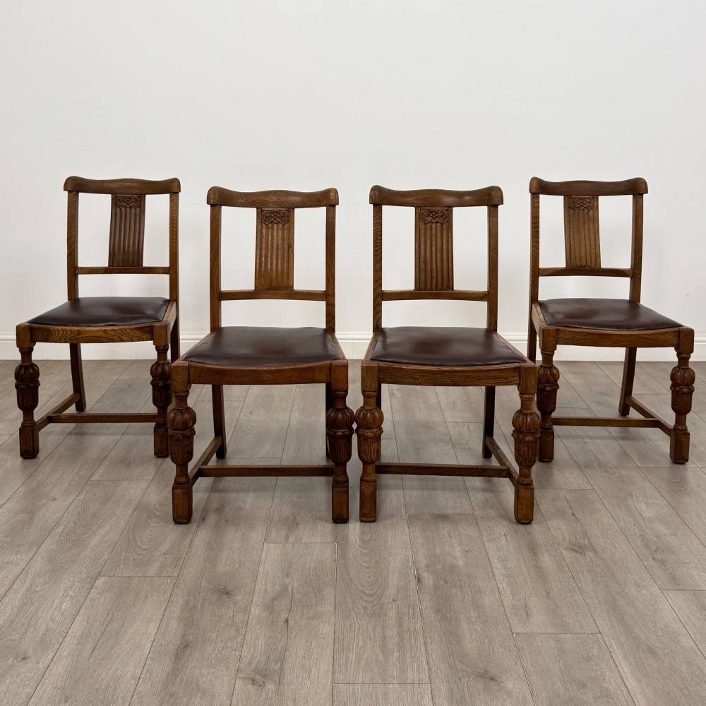 An early twentieth-century oak dining table featuring carved frieze detailing and heavy turned legs, shown alongside four matching oak dining chairs with dark leather seats. View II