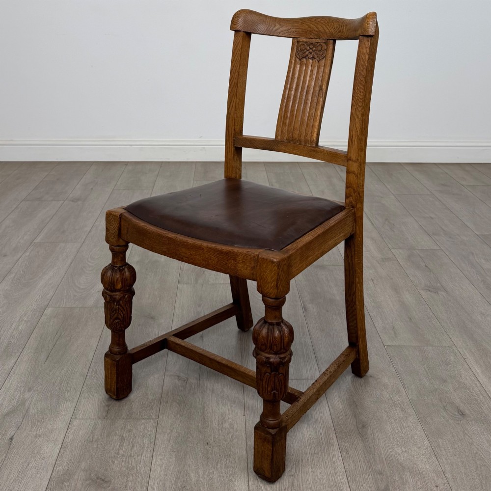 An early twentieth-century oak dining table featuring carved frieze detailing and heavy turned legs, shown alongside four matching oak dining chairs with dark leather seats. View III