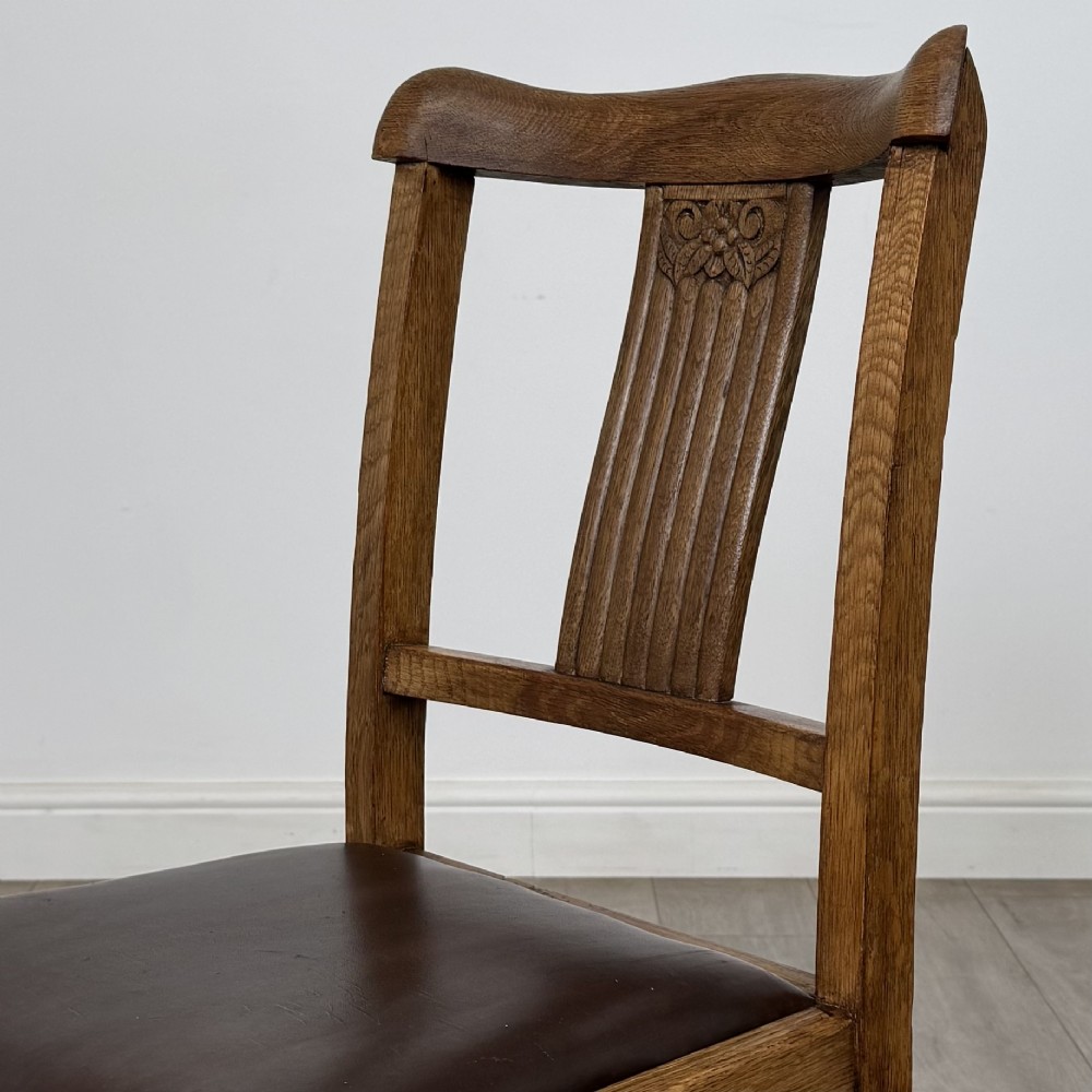 An early twentieth-century oak dining table featuring carved frieze detailing and heavy turned legs, shown alongside four matching oak dining chairs with dark leather seats. View V