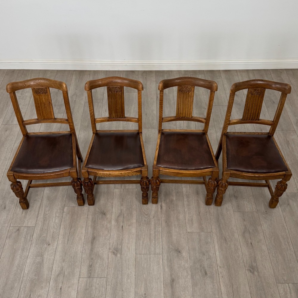 An early twentieth-century oak dining table featuring carved frieze detailing and heavy turned legs, shown alongside four matching oak dining chairs with dark leather seats. View VII