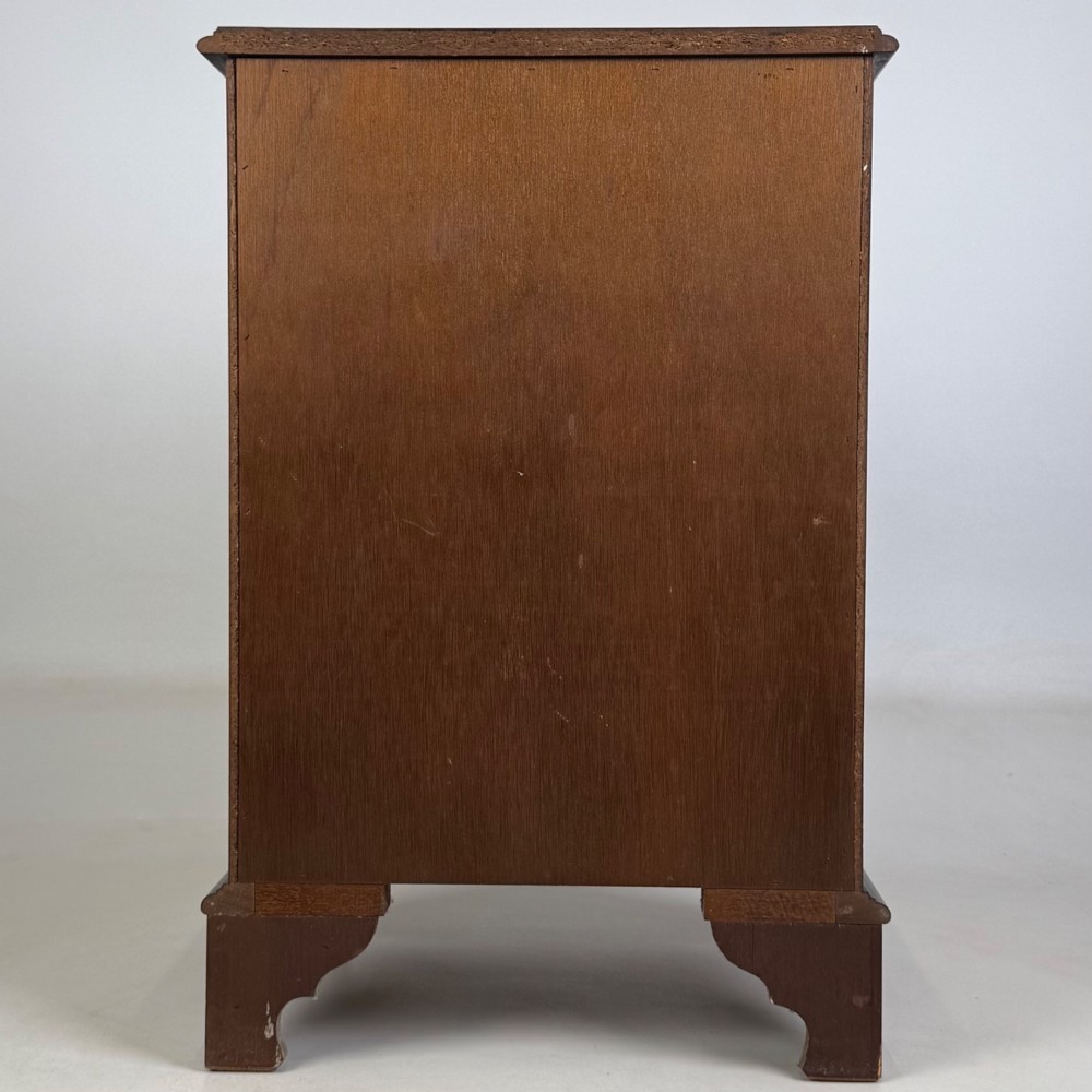 A small mahogany chest of four drawers featuring flame grain veneer fronts, circular brass ring handles, and bracket feet. View V