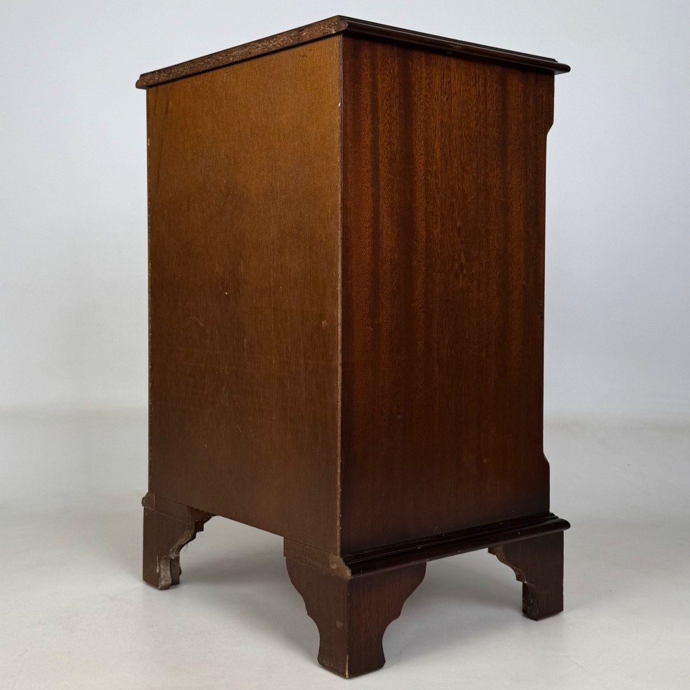 A small mahogany chest of four drawers featuring flame grain veneer fronts, circular brass ring handles, and bracket feet. View VI