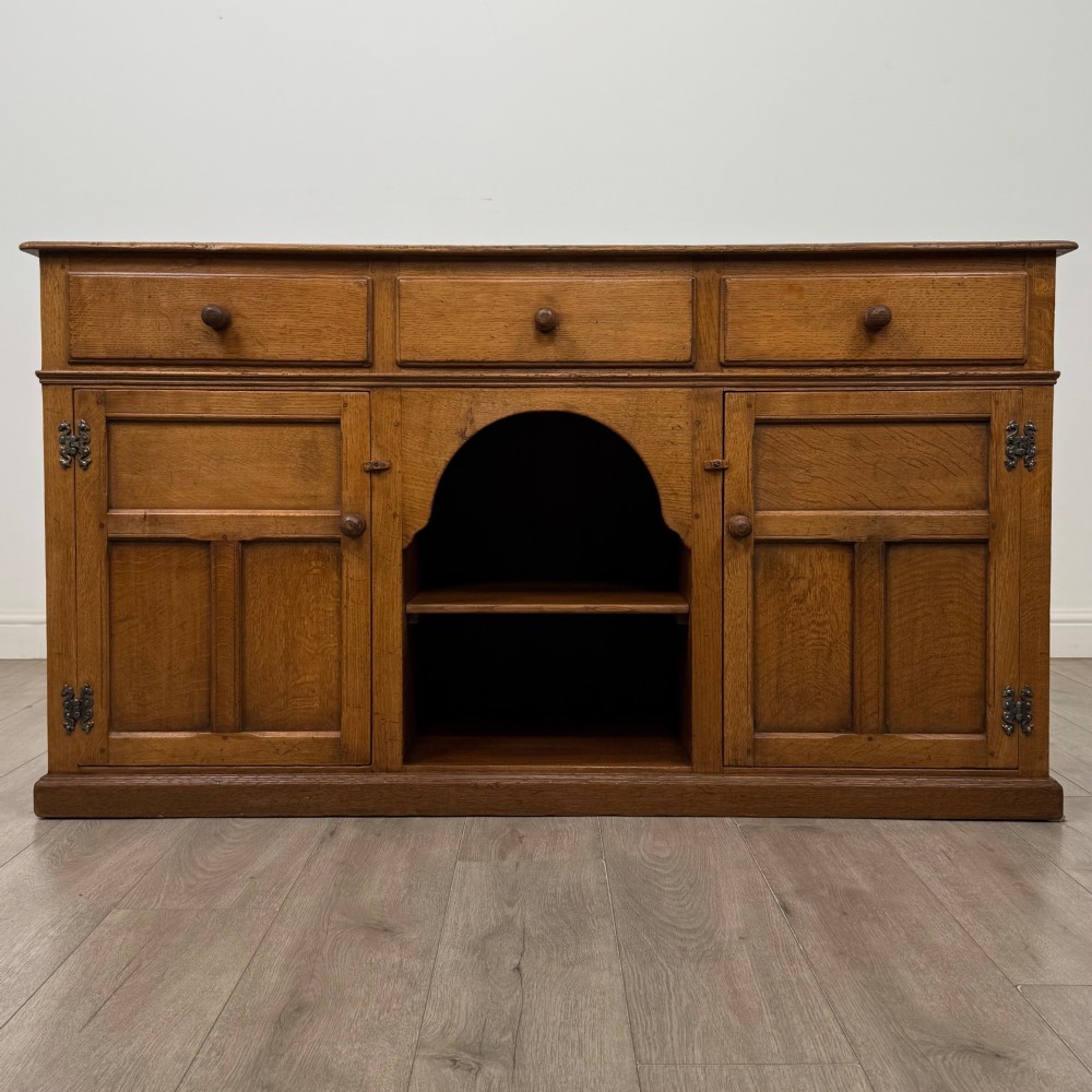 An antique golden oak dresser base featuring three top drawers, two panelled outer doors, and a central arched open recess.