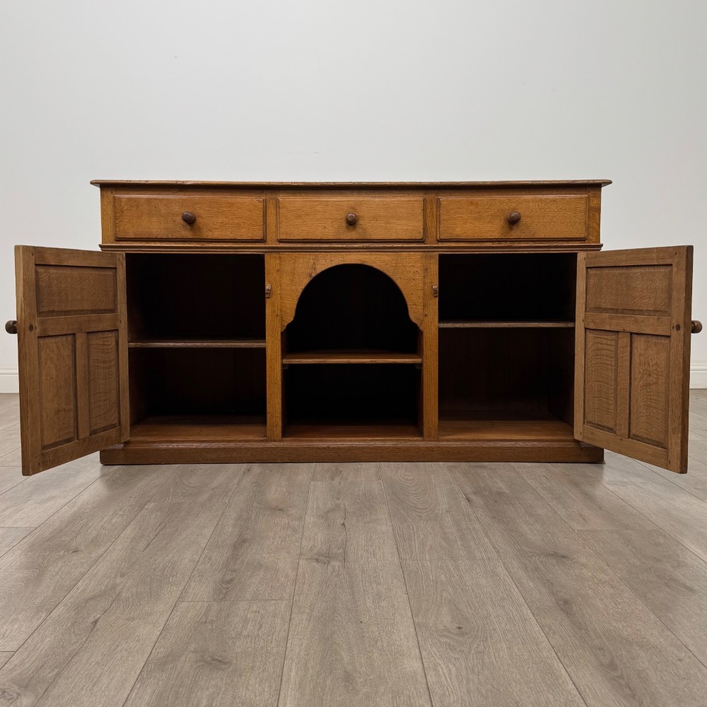 An antique golden oak dresser base featuring three top drawers, two panelled outer doors, and a central arched open recess. View II