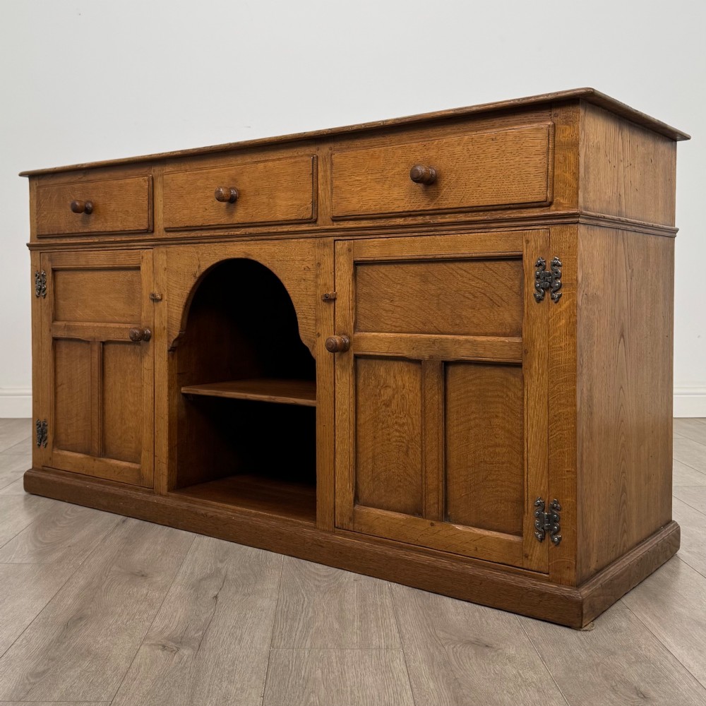 An antique golden oak dresser base featuring three top drawers, two panelled outer doors, and a central arched open recess. View III
