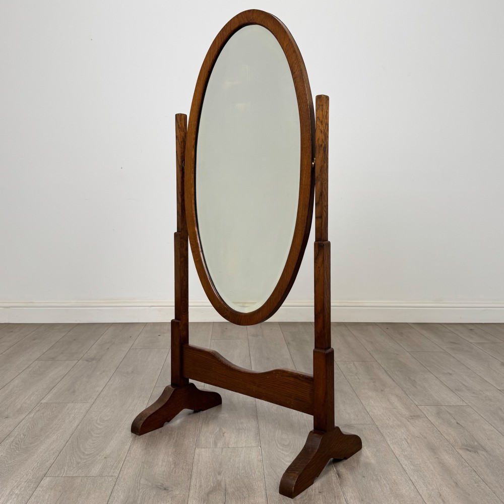 An Edwardian oak cheval mirror featuring an oval glass frame suspended between two upright posts on trestle feet.