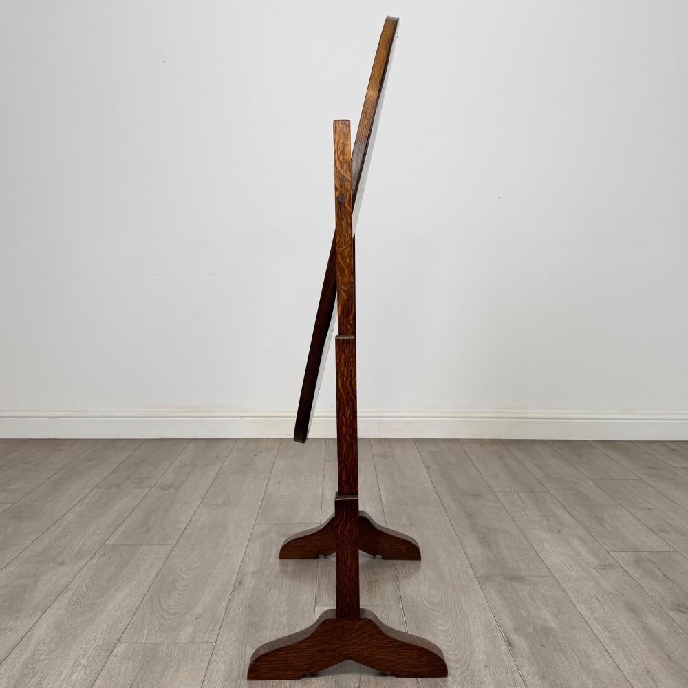 An Edwardian oak cheval mirror featuring an oval glass frame suspended between two upright posts on trestle feet. View II