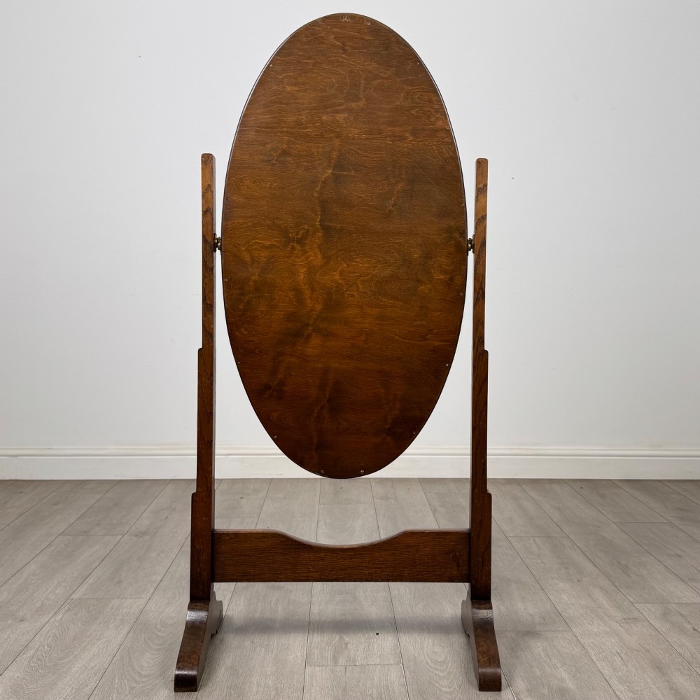 An Edwardian oak cheval mirror featuring an oval glass frame suspended between two upright posts on trestle feet. View IV