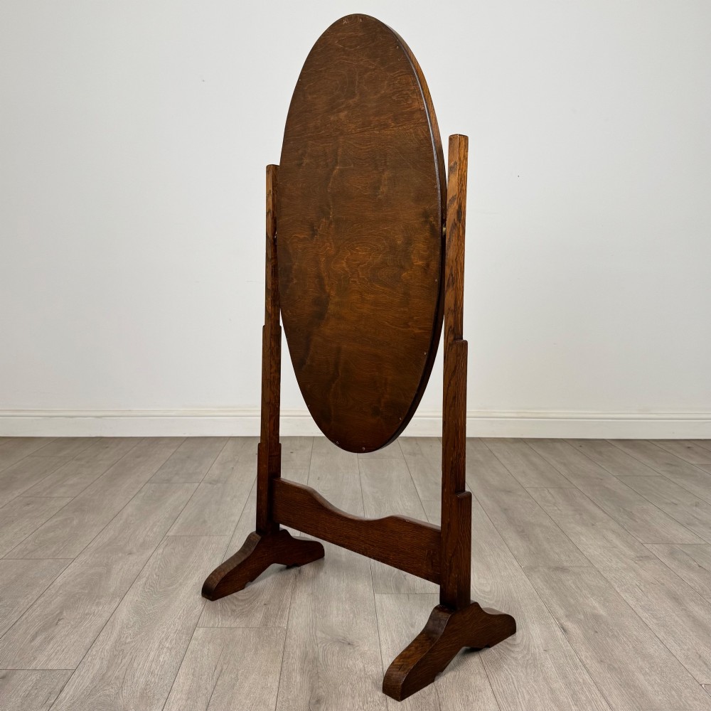 An Edwardian oak cheval mirror featuring an oval glass frame suspended between two upright posts on trestle feet. View V