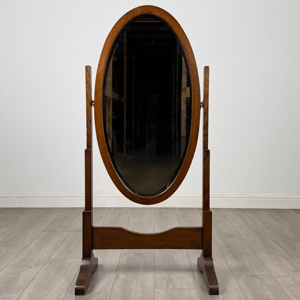An Edwardian oak cheval mirror featuring an oval glass frame suspended between two upright posts on trestle feet. View IX