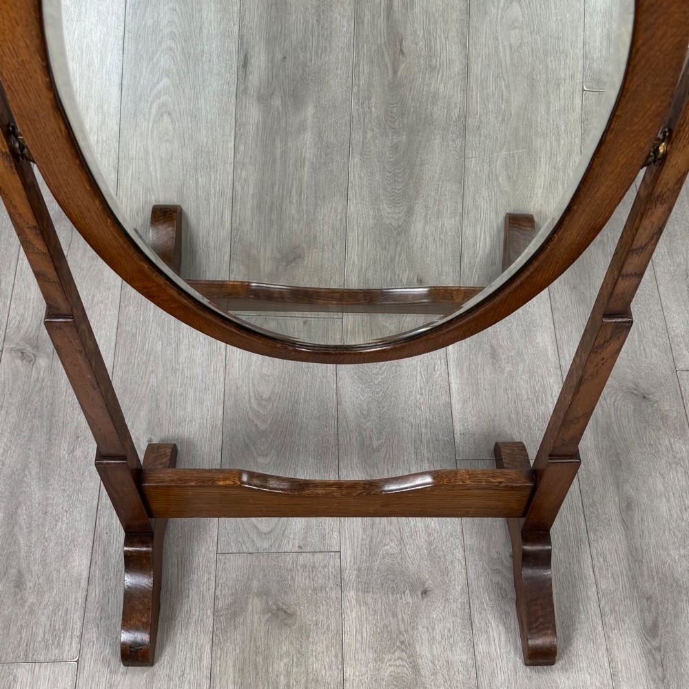 An Edwardian oak cheval mirror featuring an oval glass frame suspended between two upright posts on trestle feet. View X