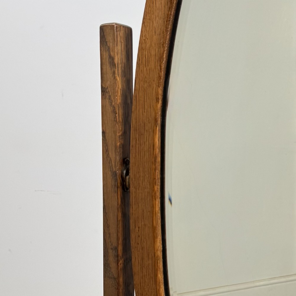 An Edwardian oak cheval mirror featuring an oval glass frame suspended between two upright posts on trestle feet. View XI